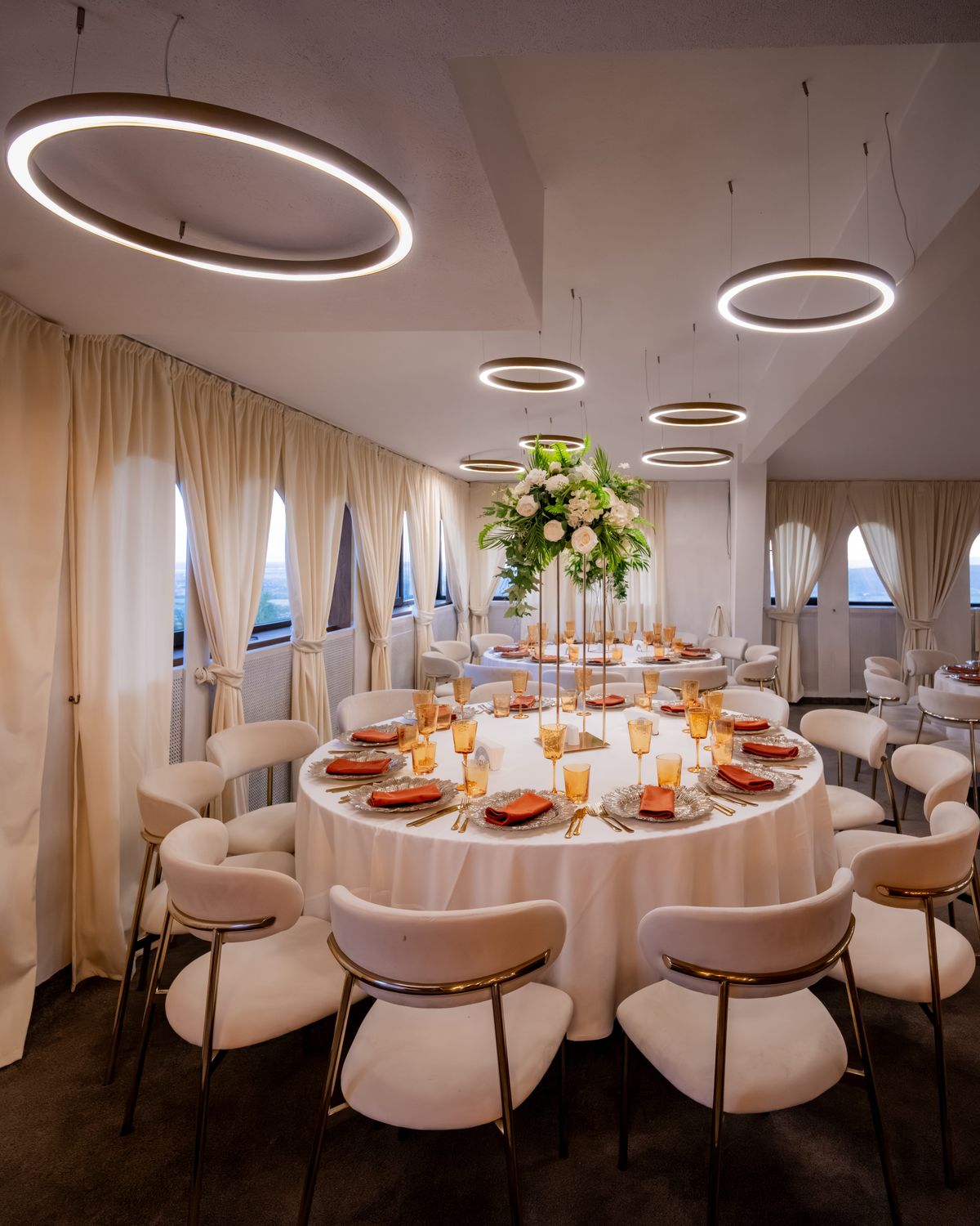 Masă rotundă aranjată într-un salon de evenimente: aranjament floral înalt, farfurii cu șervete terracotta, pahare chihlimbar și candelabre inelare deasupra