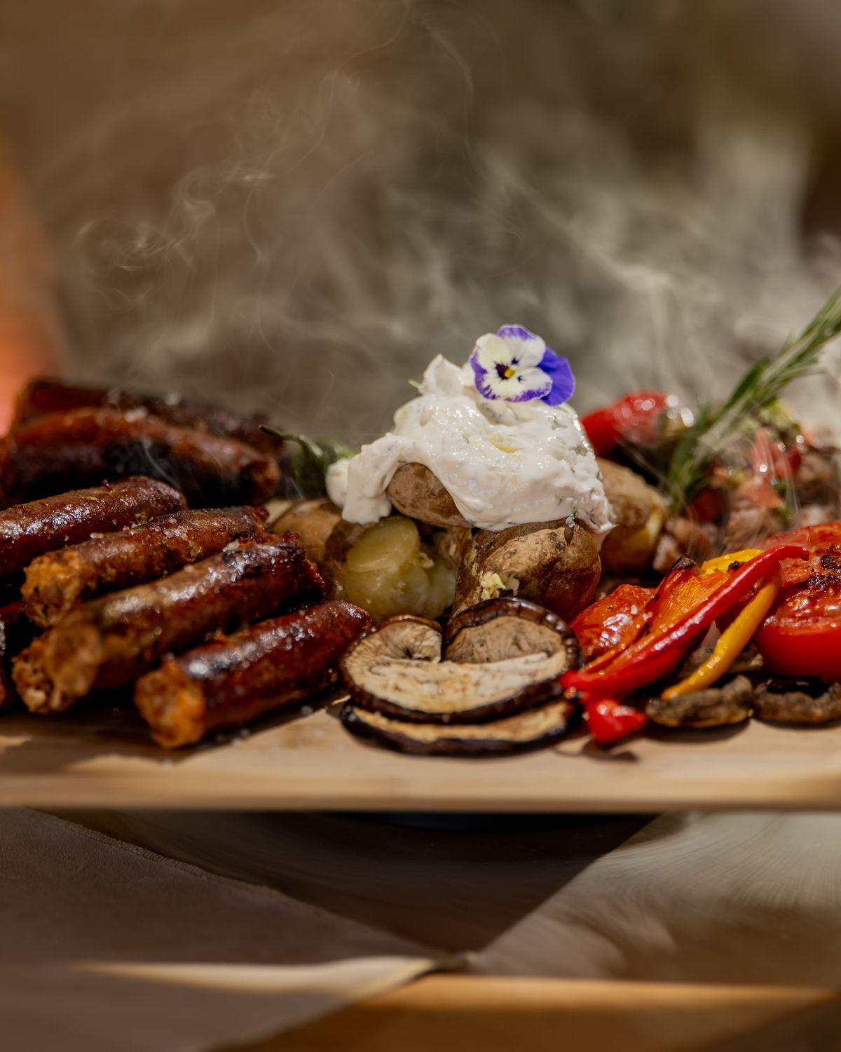 Platou de la grătar pe o planșetă de lemn: carne, ciuperci, ardei și roșii coapte, cartofi cu smântână și un fir de rozmarin
