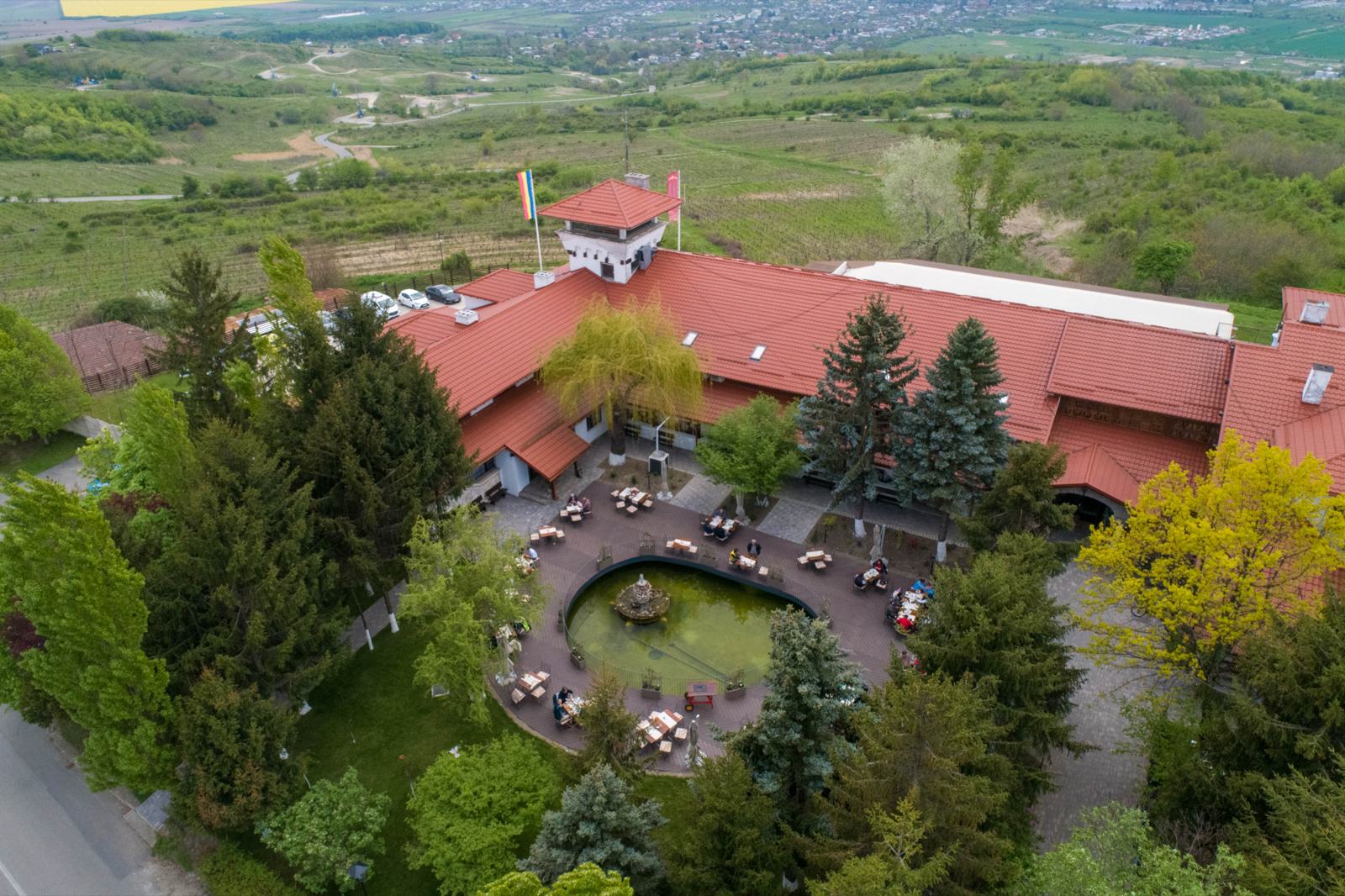 Casa Seciu văzută de sus: acoperișurile roșii și turnul, curtea cu lacul și mesele terasei, cu dealurile și viile în spate