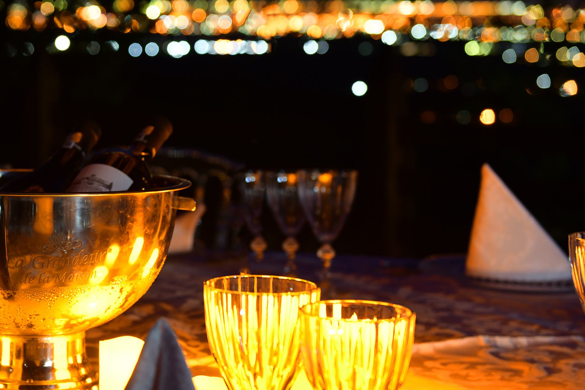 Frapieră cu sticle de vin, suporturi de lumânări și pahare pe o masă, noaptea, cu luminile orașului în vale