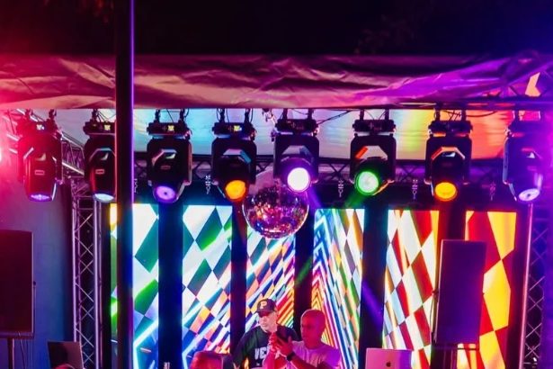Scena noaptea: lumini colorate pe grinzi, un glob disco și un perete de ecrane cu romburi, cu doi DJ la pupitru