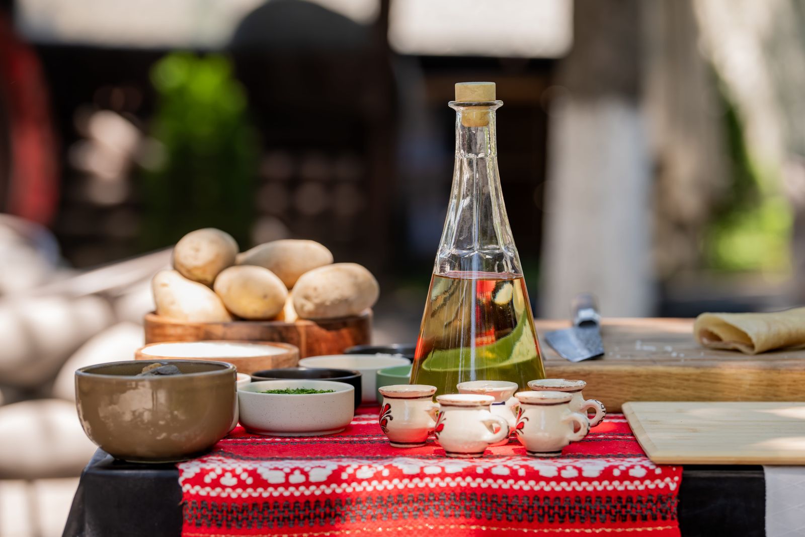 Carafă de țuică, căni de lut și castroane pe un ștergar roșu, pe masa din curte